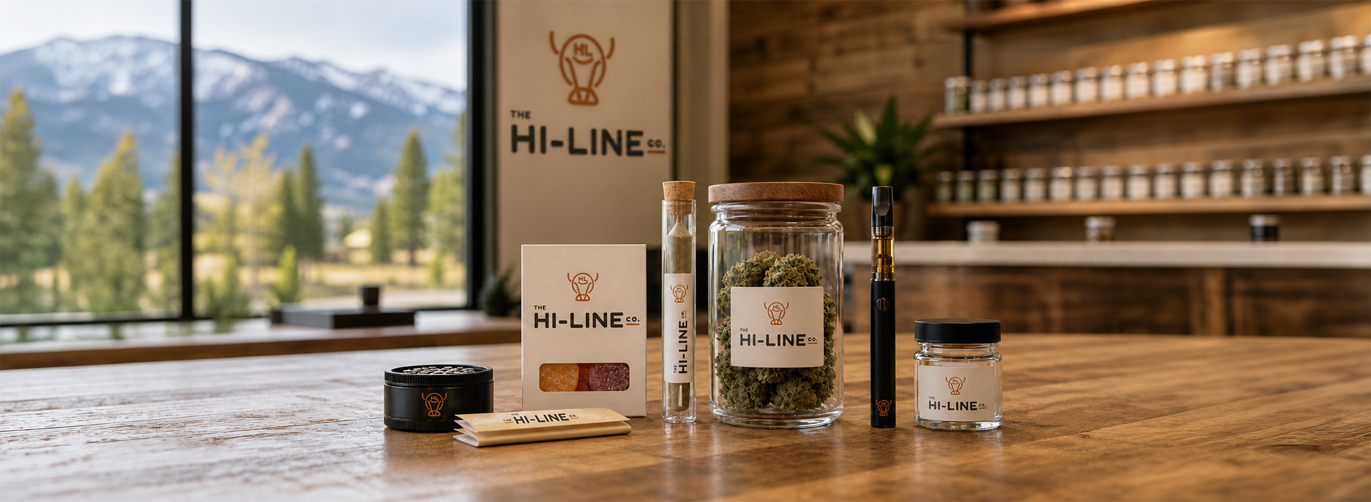 The Hi‑Line Co. cannabis products arranged on a wooden countertop, including flower jars, a vape pen, a grinder, and gummies, with a modern shop interior and mountain views in the background.