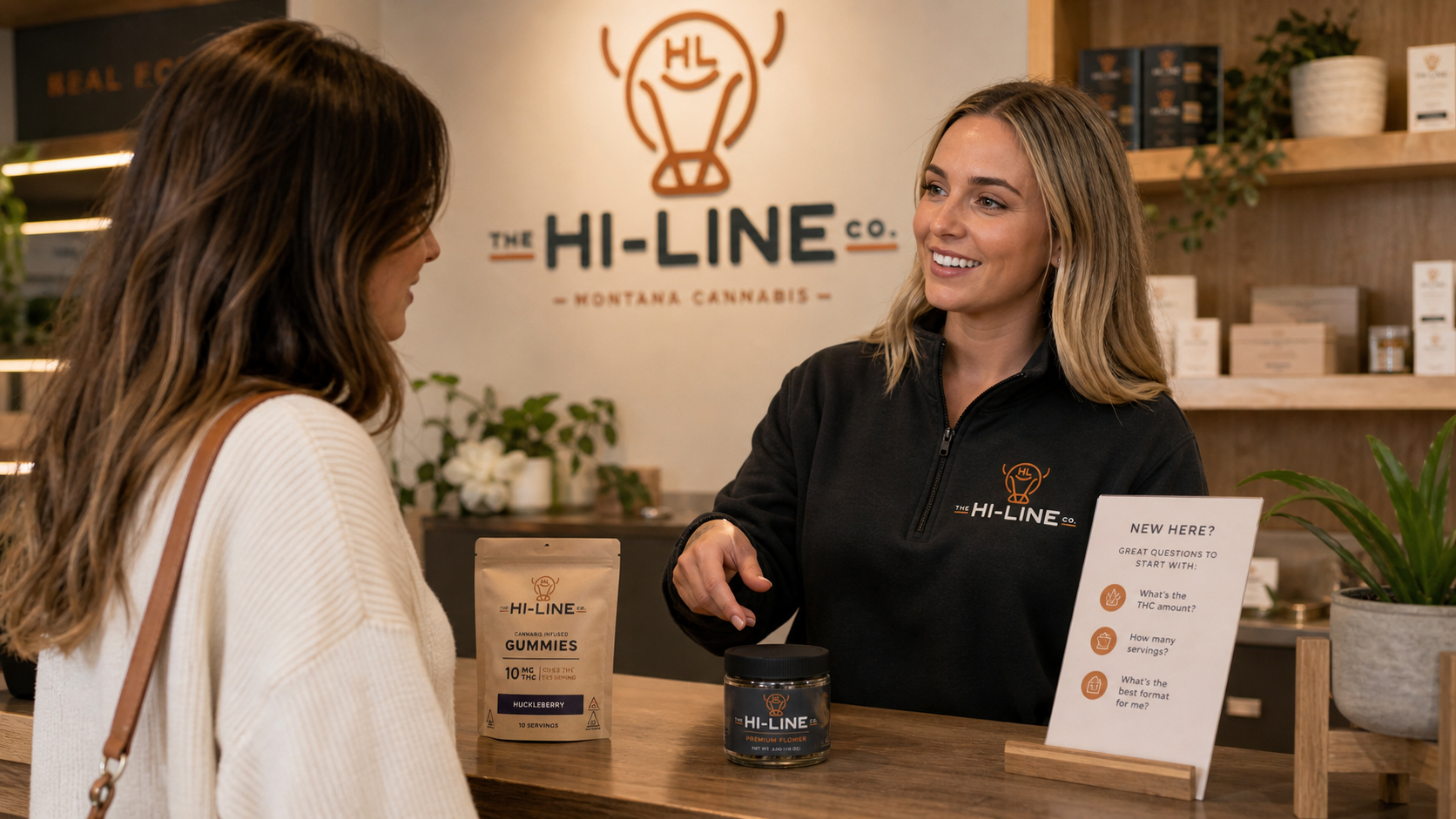A Hi‑Line Co. team member assisting a customer at the dispensary counter, with branded gummies, pre‑rolls, and flower displayed on the wood surface, and a “New Here?” educational sign and product shelves in the background.