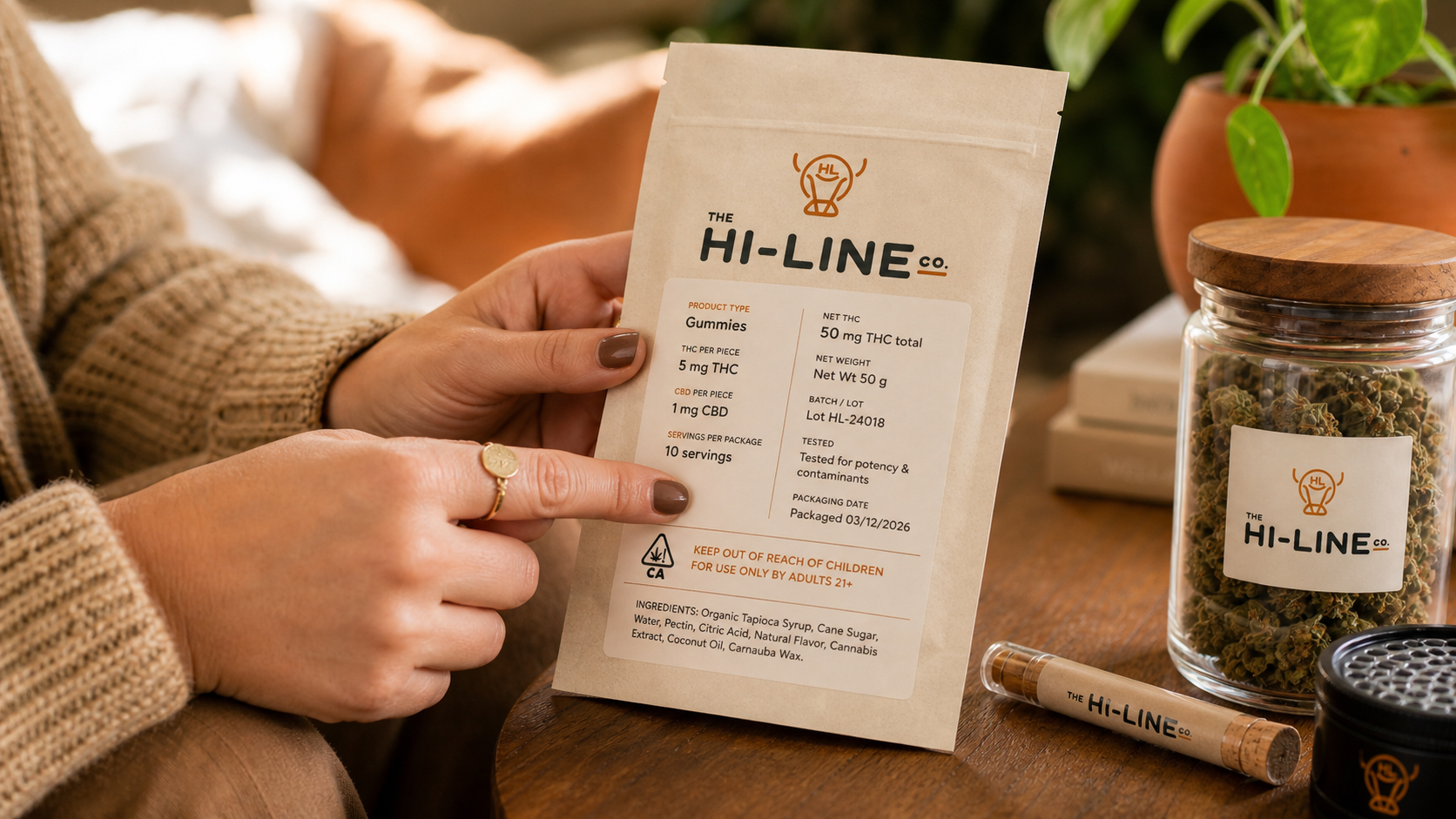 Close‑up of The Hi‑Line Co. cannabis‑infused gummies package held in hand beside a branded flower jar, grinder, and small container on a wooden table with soft indoor lighting and a plant in the background.